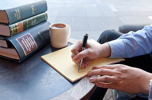 A lawyer writing notes on a legal pad beside a stack of law reports and a cup of coffee